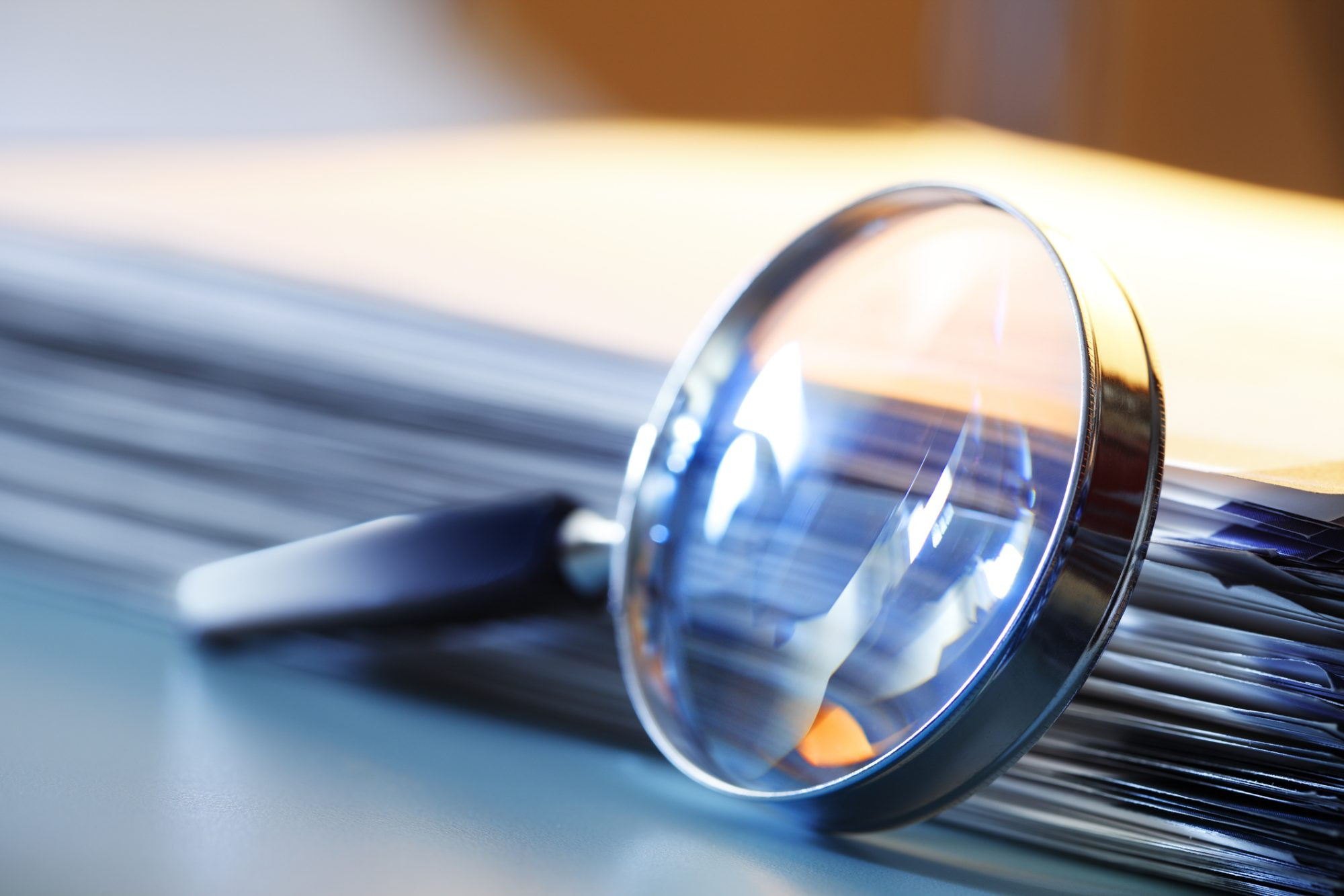 A magnifying glass leans against a thick stack of papers. Photographed with a very shallow depth of field.